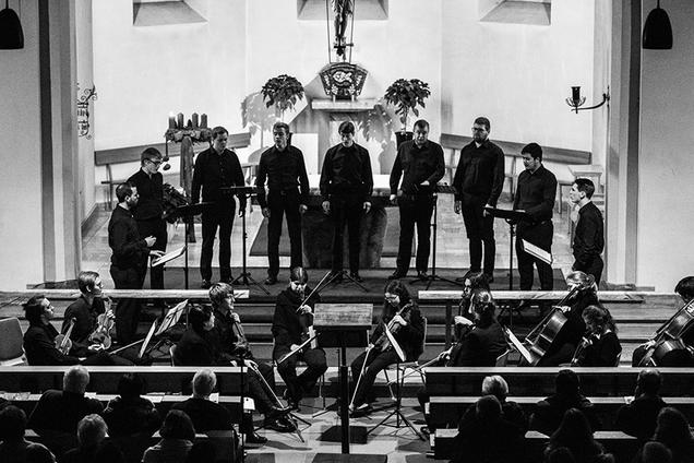 Das Foto zeigt die 9 Sänger von Principium Canti während ihres Auftritts in der Kirche St. Peter und Paul in Arzbach. Die schwarz gekleideten Sänger stehen in einem Halbkreis vor dem Altar. Vor ihnen befinden sich ihre Notenständer mit ihren Notenmappen. Vor ihnen sitzt ein Streichorchester. Am unteren Rand des Fotos sieht man ein paar der Zuschauer in den Kirchbänken sitzen.