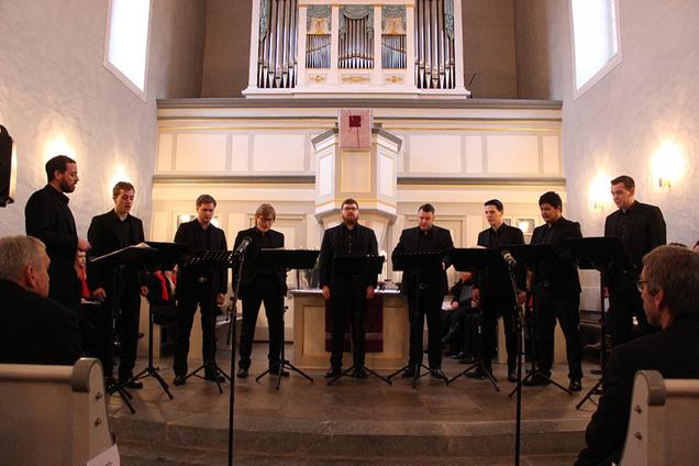 Das Foto zeigt die 9 Sänger von Principium Canti in der St.-Peter-Kirche in Diez bei Ihrem Auftritt. Die Sänger stehen vor dem Altar und tragen schwarze Hemden und Hosen. Vor ihnen stehen jeweils schwarze Notenständer mit ihren schwarzen Notenmappen. Im Hintergrund sieht man die Orgel und die Kanzel der Kirche.