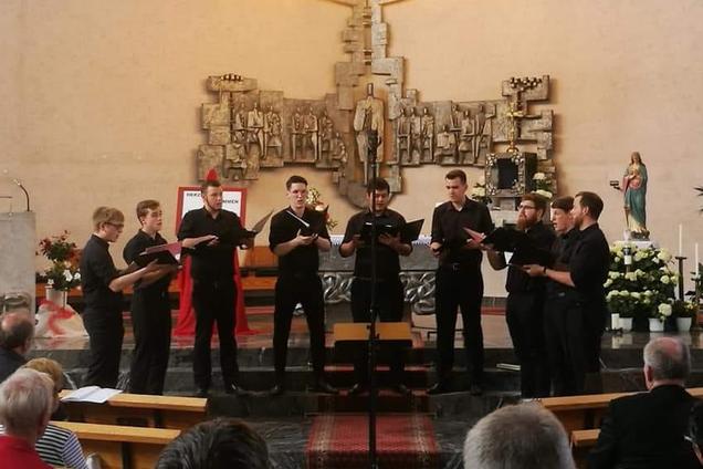 Das Foto zeigt die 9 Sänger von Principium Canti während des Chorwettbewerbs vor dem Altar der Erbacher Kirche. Die Männer tragen schwarze Hosen und Hemden und halten Ihre schwarzen Chormappen in der Hand.