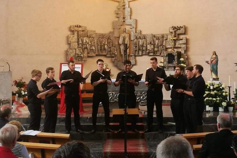 Das Foto zeigt die 9 Sänger von Principium Canti während des Chorwettbewerbs vor dem Altar der Erbacher Kirche. Die Männer tragen schwarze Hosen und Hemden und halten Ihre schwarzen Chormappen in der Hand.
