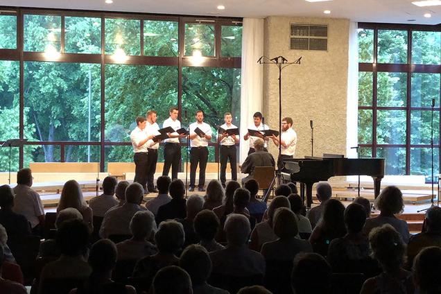 Das Foto zeigt die 7 Sänger von Principium Canti auf der Bühne während ihres Konzert im Dorfgemeinschaftshaus in Lindenholzhausen. Die Männer tragen schwarze Hosen und weiße Hemden. In der Hand halten Sie ihre schwarzen Notenmappen. Am unteren Rand des Bildes sieht man im dunkeln einen Teil der Zuschauer.