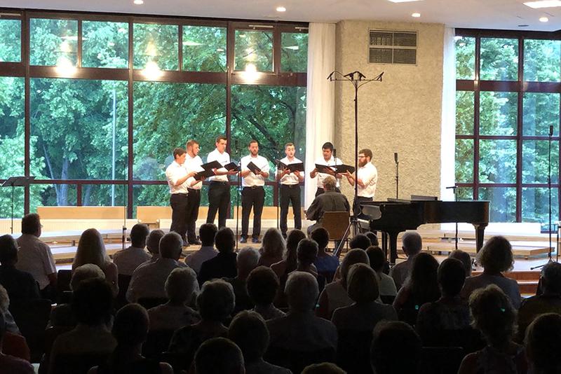 Das Foto zeigt die 7 Sänger von Principium Canti auf der Bühne während ihres Konzert im Dorfgemeinschaftshaus in Lindenholzhausen. Die Männer tragen schwarze Hosen und weiße Hemden. In der Hand halten Sie ihre schwarzen Notenmappen. Am unteren Rand des Bildes sieht man im dunkeln einen Teil der Zuschauer.
