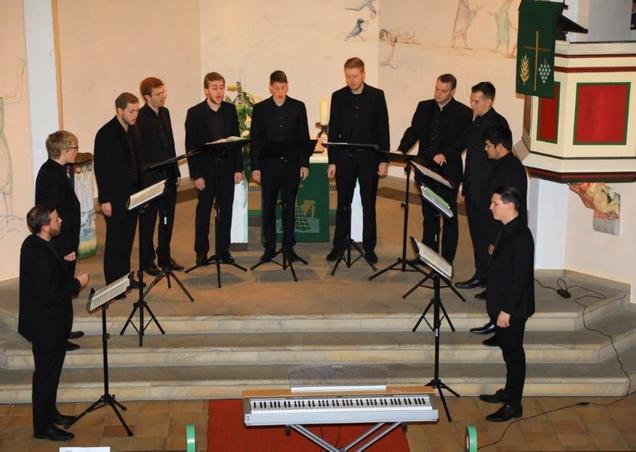 Das Foto zeigt die 11 Sänger von Principium Canti in der Fleisbacher Kirche bei ihrem Auftritt. Sie stehen vor dem Altar in ihren schwarzen Anzügen mit schwarzen Hemden. Vor ihnen steht jeweils ein schwarzer Notenständer mit ihren Notenmappen.