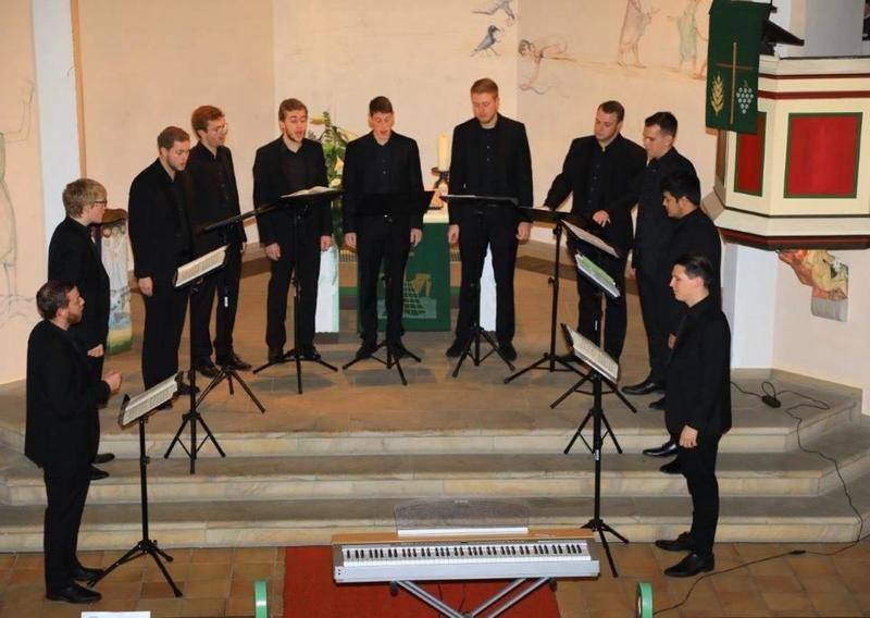 Das Foto zeigt die 11 Sänger von Principium Canti in der Fleisbacher Kirche bei ihrem Auftritt. Sie stehen vor dem Altar in ihren schwarzen Anzügen mit schwarzen Hemden. Vor ihnen steht jeweils ein schwarzer Notenständer mit ihren Notenmappen.