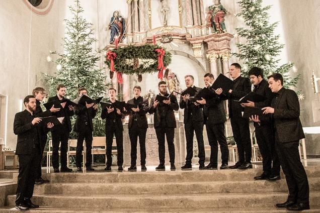 Das Foto zeigt die 12 Sänger von Principium Canti in der Kirche in Oberbrechen während ihres Auftritts. Die Sänger tragen ein schwarzes Hemd und einen schwarzen Anzug und halten Notenmappen während sie singen. Sie stehen vor dem Altar der Kirche. Im Hintergrund ist der Hochaltar zu sehen, welcher von zwei Weihnachtsbäumen eingerahmt wird. Über dem Altar hängt ein großer Adventskranz mit 4 großen roten Kerzen.