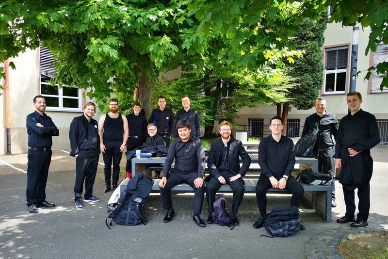 Das Foto zeigt die 12 Sänger von Principium Canti, die gerade eine Pause auf dem Schulhof der Marienschule Limburg machen, bevor sie am Limburger Chorwettbewerb in der Limburger Stadthalle teilnehmen. Alle Männer sind schwarz gekleidet. Manche tragen bereits ihr schwarzes Hemd, manche ihr Sacko und einer trägt ein schwarzes Unterhemd. Die Hälfte der Männer sitzt auf einer Bank an einen Tisch, während die andere Hälft hinter dem Tisch steht. Auch sieht man die Rucksäcke der Männer am Tisch angelehnt stehen, in denen Getränke und Notenmappen verstaut sind. Im Hintergrund ist das Gebäude der Marienschule Limburg und einige Bäume zu sehen.