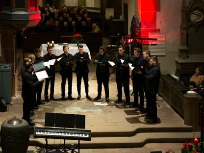 Das Foto zeigt die 12 Sänger von Principium Canti, während ihres Auftrittes in der evangelischen Stadtkirche in Herborn. Die Sänger sind gekleidet mit einem schwarzen Hemd und einem schwarzen Anzug. Sie halten ihre Notenmappen in den Händen und singen ein Lied.