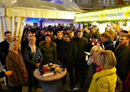 Ein Foto von Principium Canti auf dem Weihnachtsmarkt in Herborn. Die Sänger von Principium Canti stehen gemeinsam mit einer Gruppe von Frauen um einen Stehtisch mit Glühweintassen und singen ein Lied.