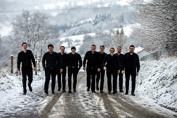 Das Bild zeigt die 9 Sänger von Principium Canti, wie sie in ihren schwarzen Hosen und Hemden eine mit Schnee bedeckte Straße hinauf laufen. Im Hintergrund ist eine weiße Schneelandschaft zu erkennen.
