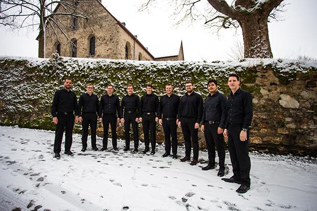 Das Foto zeigt die 9 Sänger von Principium Canti in einem Halbkreis stehend vor einer bewachsenen und beschneiten Steinmauer. Die Männer tragen schwarze Hosen und Hemden. Im Hintergrund sieht man die Kirche St. Peter und Paul in Arzbach, sowie einen Baum.